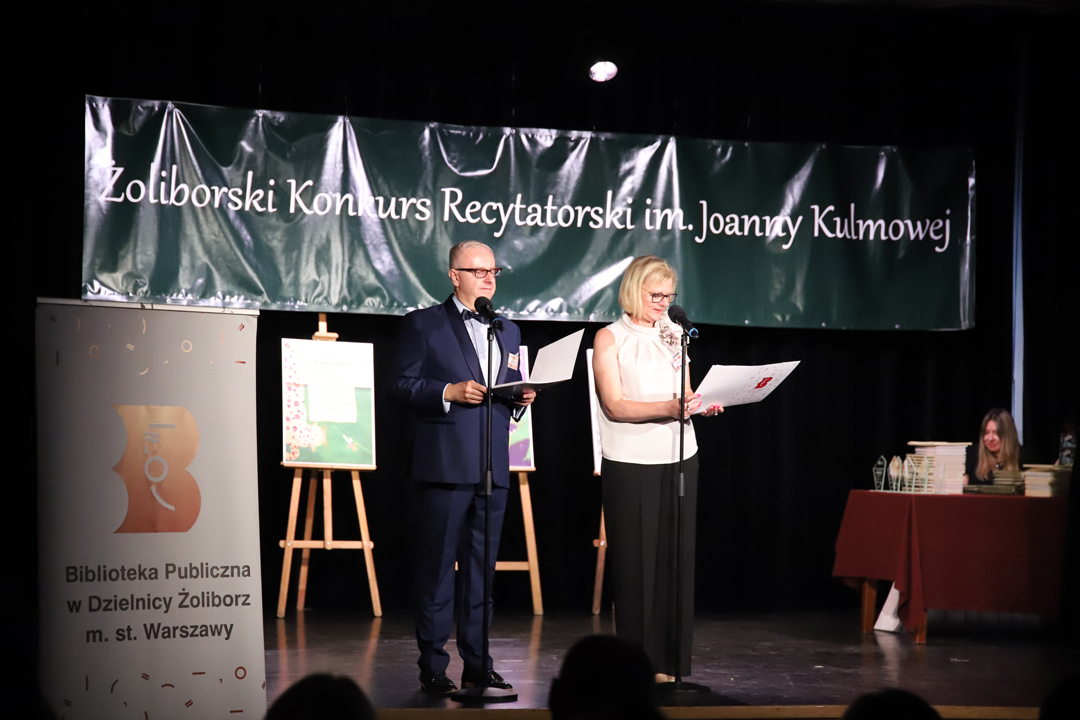 Zdjęcie z II Żoliborskiego Konkursu Recytatorskiego im. Joanny Kulmowej zorganizowanego przez Bibliotekę Publiczną w Dzielnicy Żoliborz, Społeczny Dom Kultury WSM oraz Fundację Joanny i Jana Kulmów. Dyrektor Społecznego Domu Kultury WSM oraz Biblioteki Żoliborskiej przemawiają na scenie Społecznego DOmu Kultury, w tle baner z napisem Żoliborski Konkurs Recytatorski im. Joanny Kulmowej oraz sztalugi z fragmentami utworów Joanny Kulmowej.