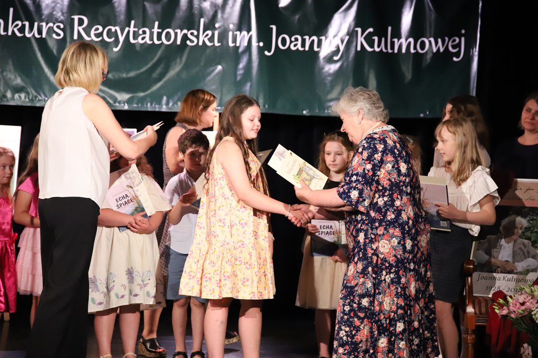 Zdjęcie z II Żoliborskiego Konkursu Recytatorskiego im. Joanny Kulmowej zorganizowanego przez Bibliotekę Publiczną w Dzielnicy Żoliborz, Społeczny Dom Kultury WSM oraz Fundację Joanny i Jana Kulmów. Laureaci konkursu - dzieci, stoją na scenie z wręczonymi nagrodami i dyplomami wraz z członkinią jury. W tle napis Żoliborski Konkurs Recytatorski im. Joanny Kulmowej oraz sztalugi z fragmentami utworów Joanny Kulmowej.