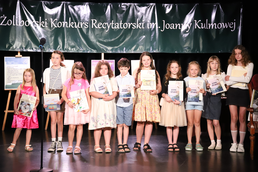 Zdjęcie z II Żoliborskiego Konkursu Recytatorskiego im. Joanny Kulmowej zorganizowanego przez Bibliotekę Publiczną w Dzielnicy Żoliborz, Społeczny Dom Kultury WSM oraz Fundację Joanny i Jana Kulmów. Laureaci konkursu - dzieci, stoją na scenie z wręczonymi nagrodami i dyplomami. W tle napis Żoliborski Konkurs Recytatorski im. Joanny Kulmowej oraz sztalugi z fragmentami utworów Joanny Kulmowej.