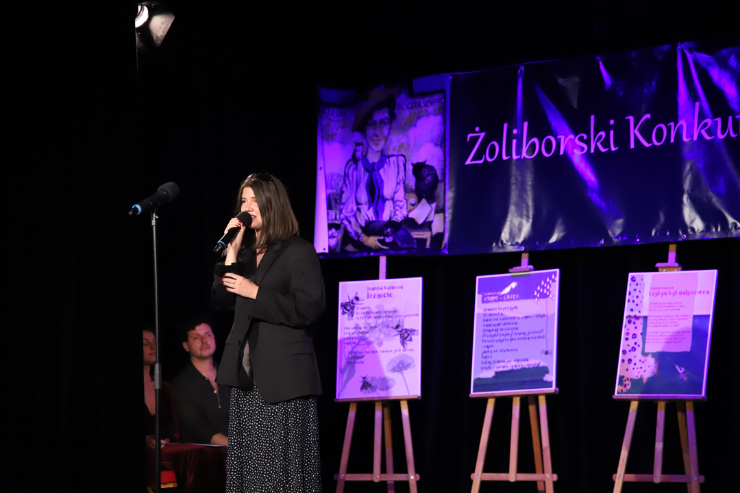Zdjęcie z II Żoliborskiego Konkursu Recytatorskiego im. Joanny Kulmowej zorganizowanego przez Bibliotekę Publiczną w Dzielnicy Żoliborz, Społeczny Dom Kultury WSM oraz Fundację Joanny i Jana Kulmów. Występ Emili Sentkowskiej śpiewającej do mikrofonu, w tle baner z napisem Żoliborski Konkurs Recytatorski im. Joanny Kulmowej oraz sztalugi z fragmentami utworów Joanny Kulmowej.
