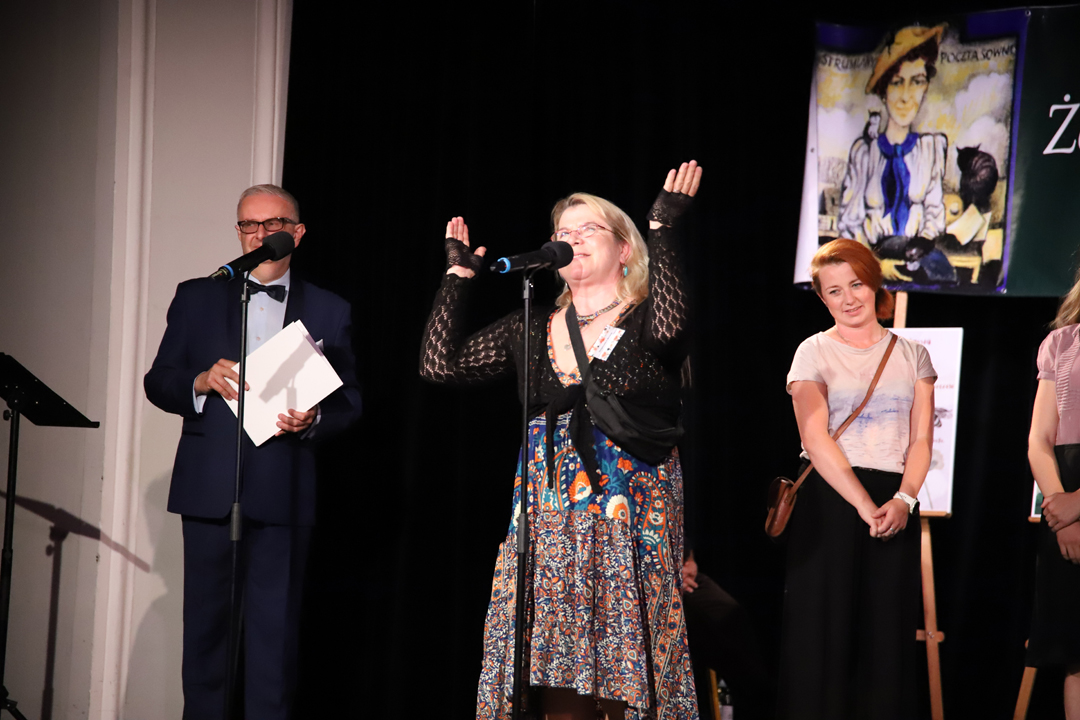 Zdjęcie z II Żoliborskiego Konkursu Recytatorskiego im. Joanny Kulmowej zorganizowanego przez Bibliotekę Publiczną w Dzielnicy Żoliborz, Społeczny Dom Kultury WSM oraz Fundację Joanny i Jana Kulmów. Małgorzata J Berwid na scenie przemawia do mikrofonu przy zgromadzonych laureatach konkursu oraz prowadzącym spotkanie Dyrektorem Społecznego Domu Kultury WSm. W tle napis Żoliborski Konkurs Recytatorski im. Joanny Kulmowej oraz sztalugi z fragmentami utworów Joanny Kulmowej.