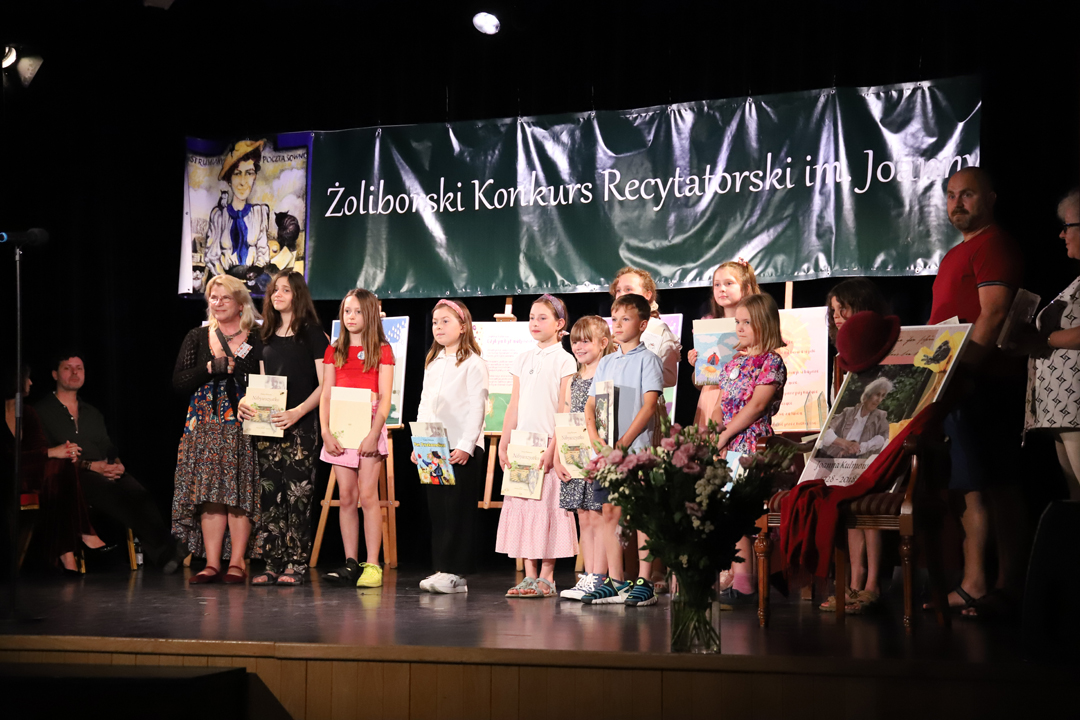 Zdjęcie z II Żoliborskiego Konkursu Recytatorskiego im. Joanny Kulmowej zorganizowanego przez Bibliotekę Publiczną w Dzielnicy Żoliborz, Społeczny Dom Kultury WSM oraz Fundację Joanny i Jana Kulmów. Laureaci konkursu - dzieci, stoją na scenie z wręczonymi nagrodami i dyplomami wraz z członkinią jury. W tle napis Żoliborski Konkurs Recytatorski im. Joanny Kulmowej oraz sztalugi z fragmentami utworów Joanny Kulmowej.