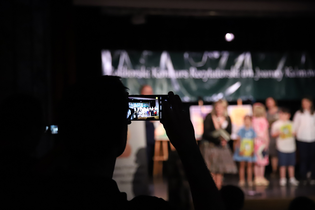Zdjęcie z II Żoliborskiego Konkursu Recytatorskiego im. Joanny Kulmowej zorganizowanego przez Bibliotekę Publiczną w Dzielnicy Żoliborz, Społeczny Dom Kultury WSM oraz Fundację Joanny i Jana Kulmów. Uczestnik spotkania robi zdjęcie telefonem komórkowym. W tle laureaci konkursu stojący na scenie oraz baner z napisem Żoliborski Konkurs Recytatorski im. Joanny Kulmowej oraz sztalugi z fragmentami utworów Joanny Kulmowej.