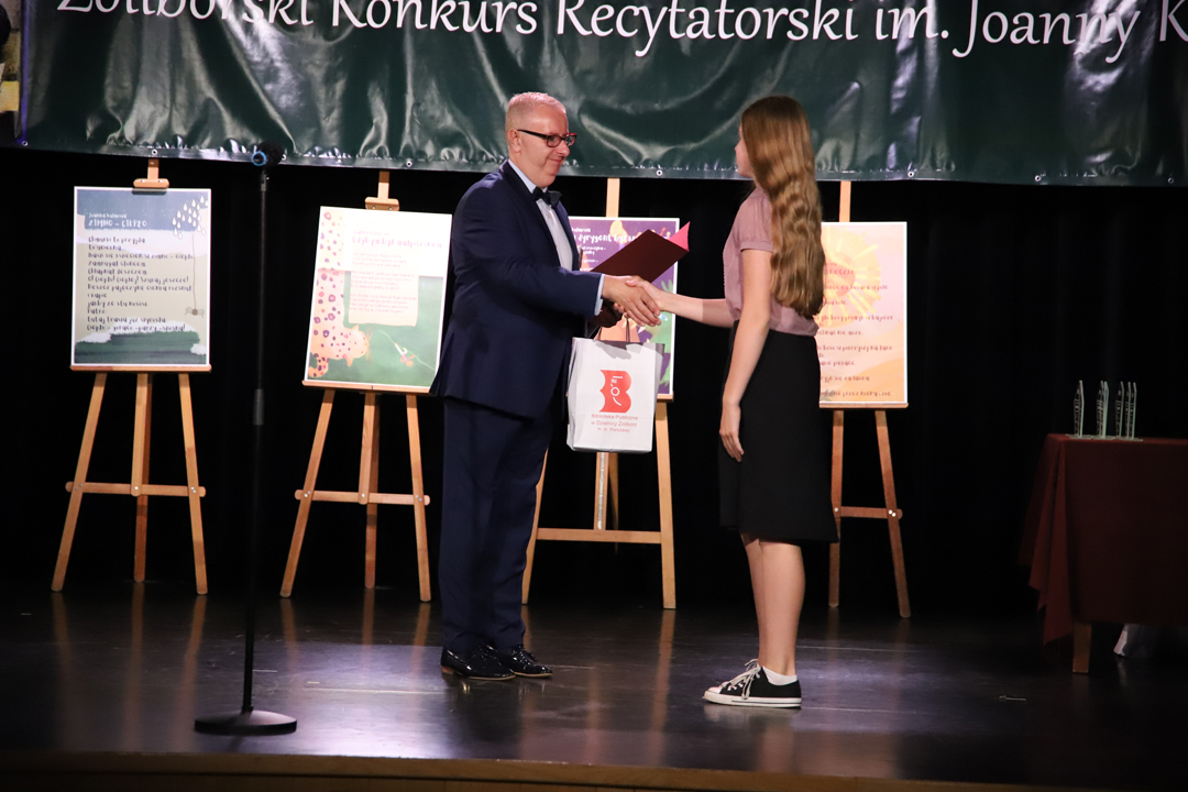 Zdjęcie z II Żoliborskiego Konkursu Recytatorskiego im. Joanny Kulmowej zorganizowanego przez Bibliotekę Publiczną w Dzielnicy Żoliborz, Społeczny Dom Kultury WSM oraz Fundację Joanny i Jana Kulmów. Członek Jury wręcza nagrody i dyplomy na scenie Społecznego Domu Kultury. W tle baner z napisem Żoliborski Konkurs Recytatorski im. Joanny Kulmowej oraz sztalugi z fragmentami utworów Joanny Kulmowej.