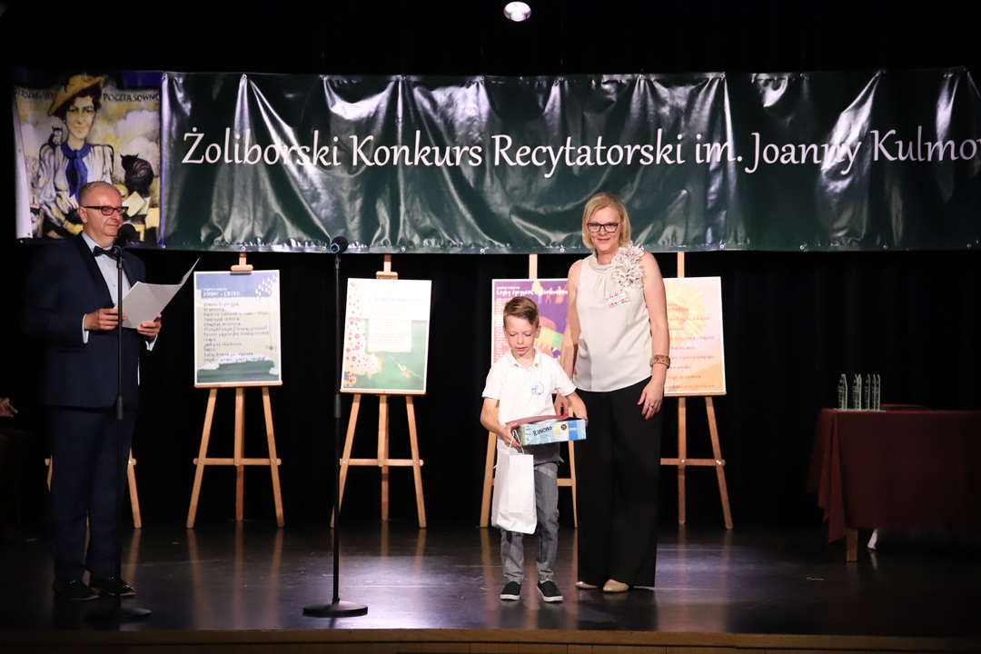 Zdjęcie z II Żoliborskiego Konkursu Recytatorskiego im. Joanny Kulmowej zorganizowanego przez Bibliotekę Publiczną w Dzielnicy Żoliborz, Społeczny Dom Kultury WSM oraz Fundację Joanny i Jana Kulmów. Członek Jury wręcza nagrody i dyplomy na scenie Społecznego Domu Kultury. W tle Dyrektor Społecznego Domu Kultury WSM oraz baner z napisem Żoliborski Konkurs Recytatorski im. Joanny Kulmowej oraz sztalugi z fragmentami utworów Joanny Kulmowej.