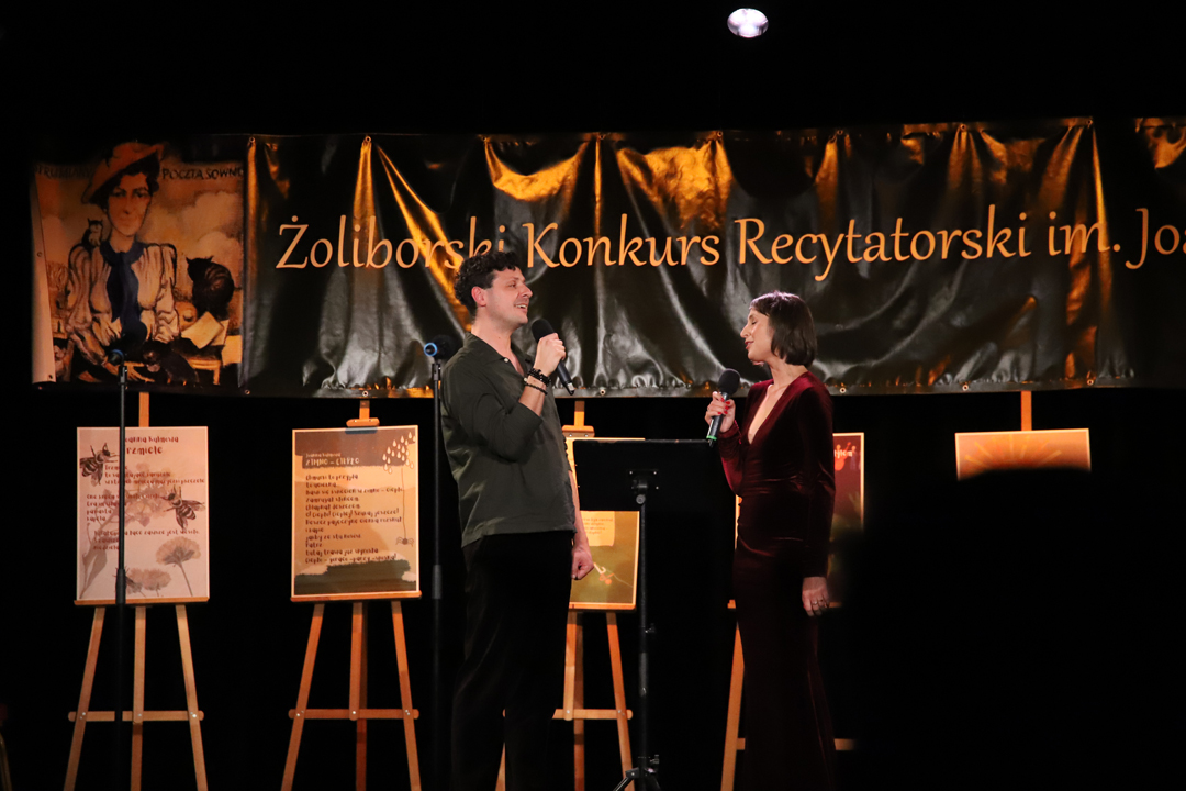 Zdjęcie z II Żoliborskiego Konkursu Recytatorskiego im. Joanny Kulmowej zorganizowanego przez Bibliotekę Publiczną w Dzielnicy Żoliborz, Społeczny Dom Kultury WSM oraz Fundację Joanny i Jana Kulmów. Artyści występujący na scenie Społecznego Domu Kultury śpiewają przy mikrofonach. W tle baner z napisem Żoliborski Konkurs Recytatorski im. Joanny Kulmowej oraz sztalugi z fragmentami utworów Joanny Kulmowej.