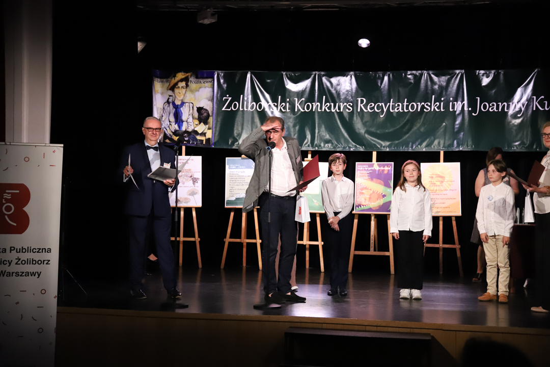 Zdjęcie z II Żoliborskiego Konkursu Recytatorskiego im. Joanny Kulmowej zorganizowanego przez Bibliotekę Publiczną w Dzielnicy Żoliborz, Społeczny Dom Kultury WSM oraz Fundację Joanny i Jana Kulmów. Członek Jury wręcza nagrody i dyplomy na scenie Społecznego Domu Kultury. W tle Dyrektor Społecznego Domu Kultury WSM oraz baner z napisem Żoliborski Konkurs Recytatorski im. Joanny Kulmowej oraz sztalugi z fragmentami utworów Joanny Kulmowej.