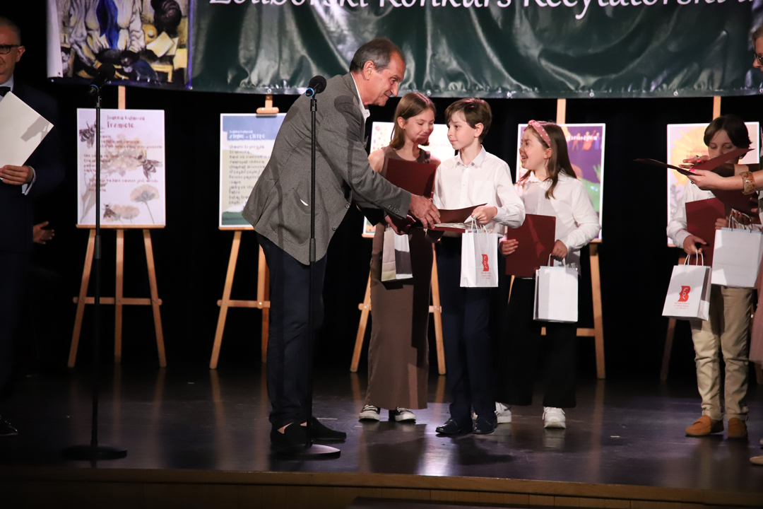 Zdjęcie z II Żoliborskiego Konkursu Recytatorskiego im. Joanny Kulmowej zorganizowanego przez Bibliotekę Publiczną w Dzielnicy Żoliborz, Społeczny Dom Kultury WSM oraz Fundację Joanny i Jana Kulmów. Członek Jury wręcza nagrody i dyplomy na scenie Społecznego Domu Kultury. W tle Dyrektor Społecznego Domu Kultury WSM oraz baner z napisem Żoliborski Konkurs Recytatorski im. Joanny Kulmowej oraz sztalugi z fragmentami utworów Joanny Kulmowej.