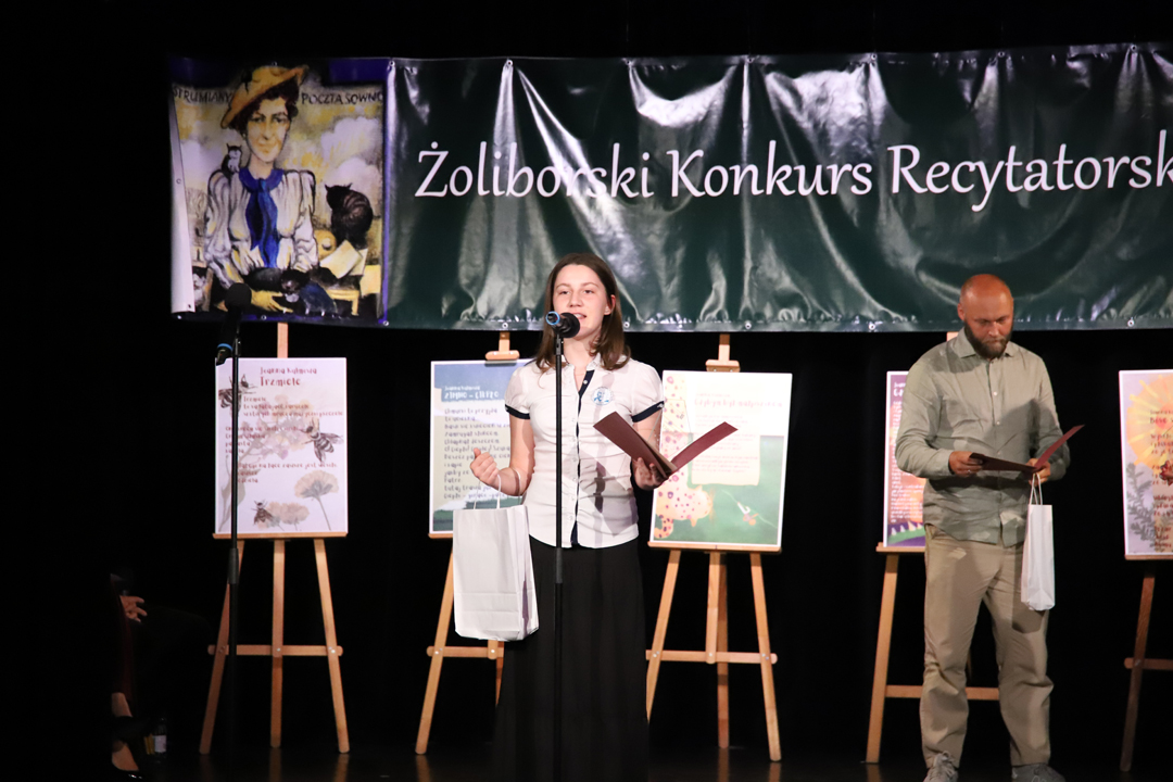 Zdjęcie z II Żoliborskiego Konkursu Recytatorskiego im. Joanny Kulmowej zorganizowanego przez Bibliotekę Publiczną w Dzielnicy Żoliborz, Społeczny Dom Kultury WSM oraz Fundację Joanny i Jana Kulmów. Występ laureatki konkursu. W tle członek jury oraz baner z napisem Żoliborski Konkurs Recytatorski im. Joanny Kulmowej oraz sztalugi z fragmentami utworów Joanny Kulmowej.