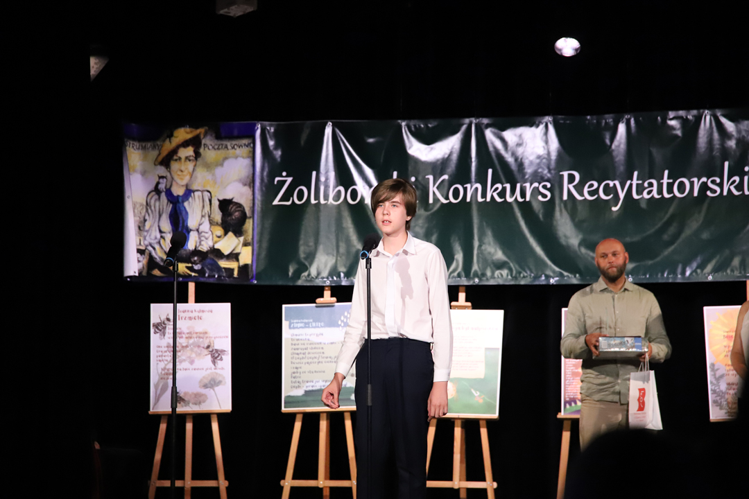 Zdjęcie z II Żoliborskiego Konkursu Recytatorskiego im. Joanny Kulmowej zorganizowanego przez Bibliotekę Publiczną w Dzielnicy Żoliborz, Społeczny Dom Kultury WSM oraz Fundację Joanny i Jana Kulmów. Występ laureatki konkursu. W tle członek jury oraz baner z napisem Żoliborski Konkurs Recytatorski im. Joanny Kulmowej oraz sztalugi z fragmentami utworów Joanny Kulmowej.