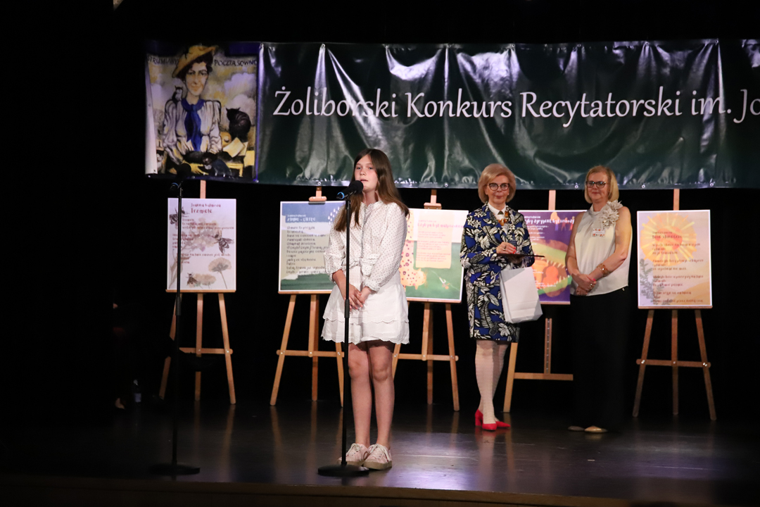 Zdjęcie z II Żoliborskiego Konkursu Recytatorskiego im. Joanny Kulmowej zorganizowanego przez Bibliotekę Publiczną w Dzielnicy Żoliborz, Społeczny Dom Kultury WSM oraz Fundację Joanny i Jana Kulmów. Występ laureatki konkursu. W tle członkinie jury oraz baner z napisem Żoliborski Konkurs Recytatorski im. Joanny Kulmowej oraz sztalugi z fragmentami utworów Joanny Kulmowej.