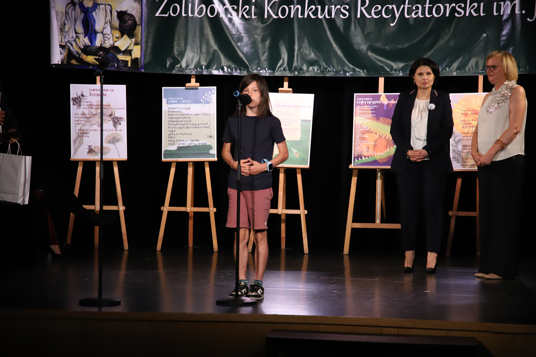 Zdjęcie z II Żoliborskiego Konkursu Recytatorskiego im. Joanny Kulmowej zorganizowanego przez Bibliotekę Publiczną w Dzielnicy Żoliborz, Społeczny Dom Kultury WSM oraz Fundację Joanny i Jana Kulmów. Występ laureatki konkursu. W tle członkinie jury oraz baner z napisem Żoliborski Konkurs Recytatorski im. Joanny Kulmowej oraz sztalugi z fragmentami utworów Joanny Kulmowej.