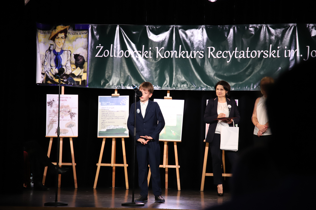 Zdjęcie z II Żoliborskiego Konkursu Recytatorskiego im. Joanny Kulmowej zorganizowanego przez Bibliotekę Publiczną w Dzielnicy Żoliborz, Społeczny Dom Kultury WSM oraz Fundację Joanny i Jana Kulmów. Występ laureatki konkursu. W tle członkinie jury oraz baner z napisem Żoliborski Konkurs Recytatorski im. Joanny Kulmowej oraz sztalugi z fragmentami utworów Joanny Kulmowej.