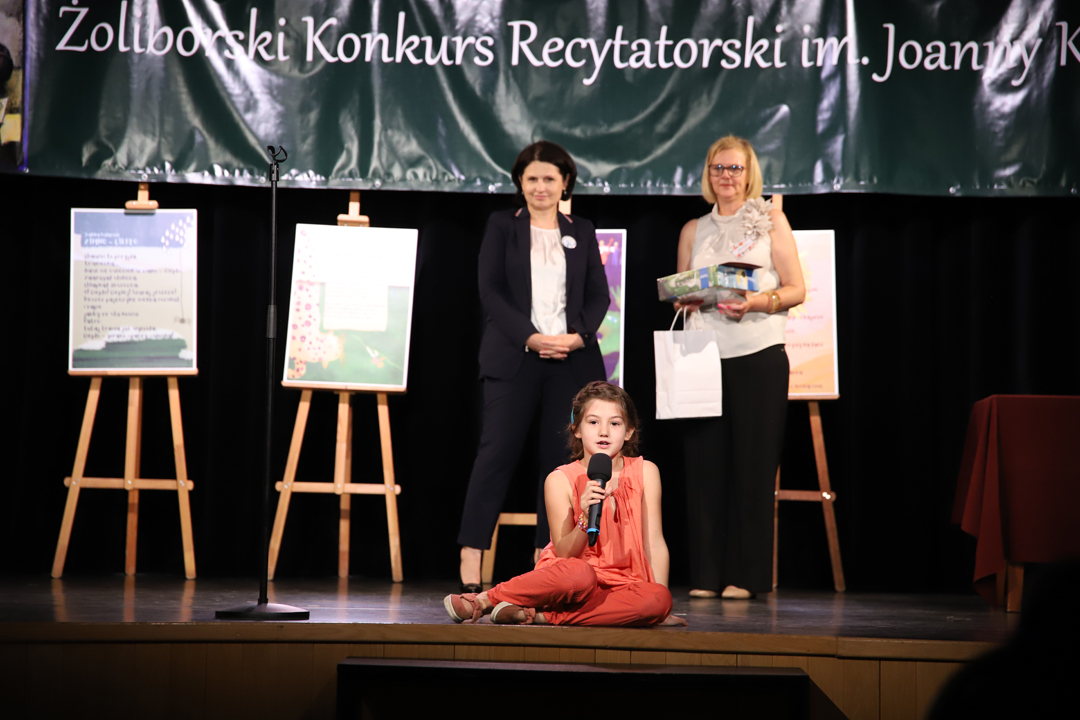 Zdjęcie z II Żoliborskiego Konkursu Recytatorskiego im. Joanny Kulmowej zorganizowanego przez Bibliotekę Publiczną w Dzielnicy Żoliborz, Społeczny Dom Kultury WSM oraz Fundację Joanny i Jana Kulmów. Dziewczynka siedząca na scenie i recytująca do mikrofonu utwór Joanny Kulmowej. W tle Burmistrzyni Żoliborza oraz Dyrektor Biblioteki stojące na scenie oraz baner z napisem Żoliborski Konkurs Recytatorski im. Joanny Kulmowej oraz sztalugi z fragmentami utworów Joanny Kulmowej.