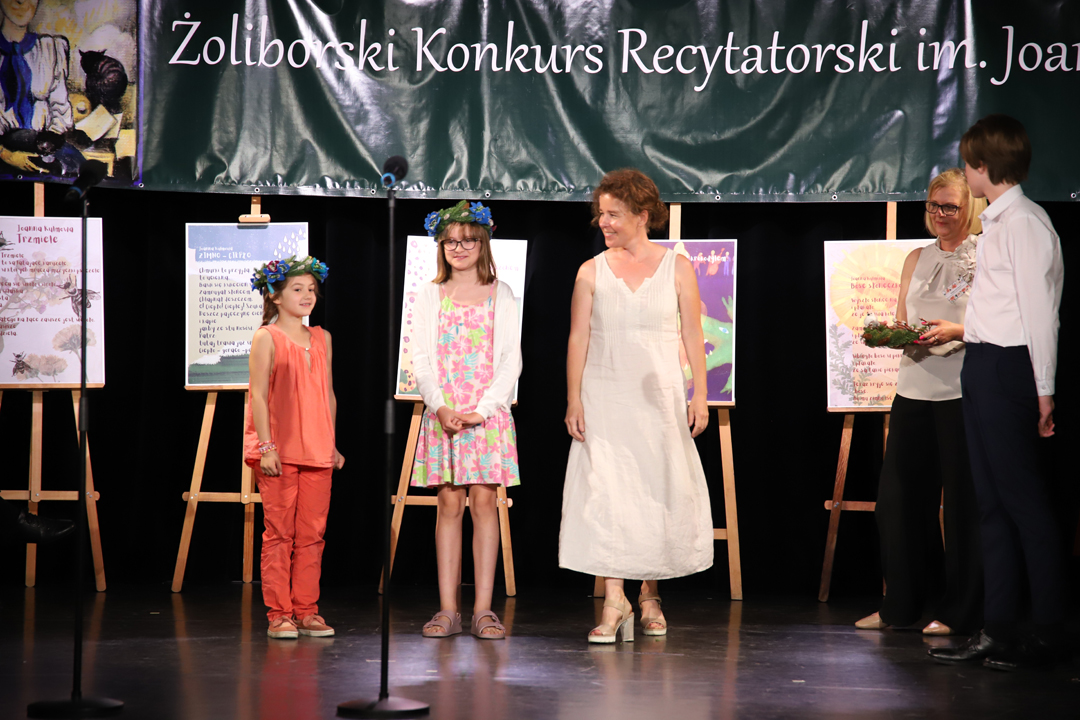 Zdjęcie z II Żoliborskiego Konkursu Recytatorskiego im. Joanny Kulmowej zorganizowanego przez Bibliotekę Publiczną w Dzielnicy Żoliborz, Społeczny Dom Kultury WSM oraz Fundację Joanny i Jana Kulmów. Członek Jury wręcza nagrody i dyplomy na scenie Społecznego Domu Kultury. W tle baner z napisem Żoliborski Konkurs Recytatorski im. Joanny Kulmowej oraz sztalugi z fragmentami utworów Joanny Kulmowej.