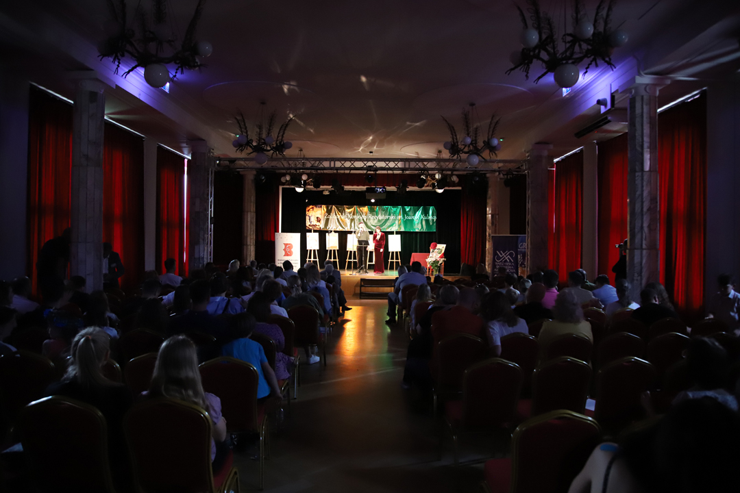 Zdjęcie z II Żoliborskiego Konkursu Recytatorskiego im. Joanny Kulmowej zorganizowanego przez Bibliotekę Publiczną w Dzielnicy Żoliborz, Społeczny Dom Kultury WSM oraz Fundację Joanny i Jana Kulmów. Widok na publiczność siedzącą przed sceną Społecznego Domu Kultury. W tle artyści występujacy na scenie oraz baner z napisem Żoliborski Konkurs Recytatorski im. Joanny Kulmowej oraz sztalugi z fragmentami utworów Joanny Kulmowej oraz rollupem Biblioteki Żoliborskiej z napisem Biblioteka Publiczna w Dzielnicy Żoliborz.