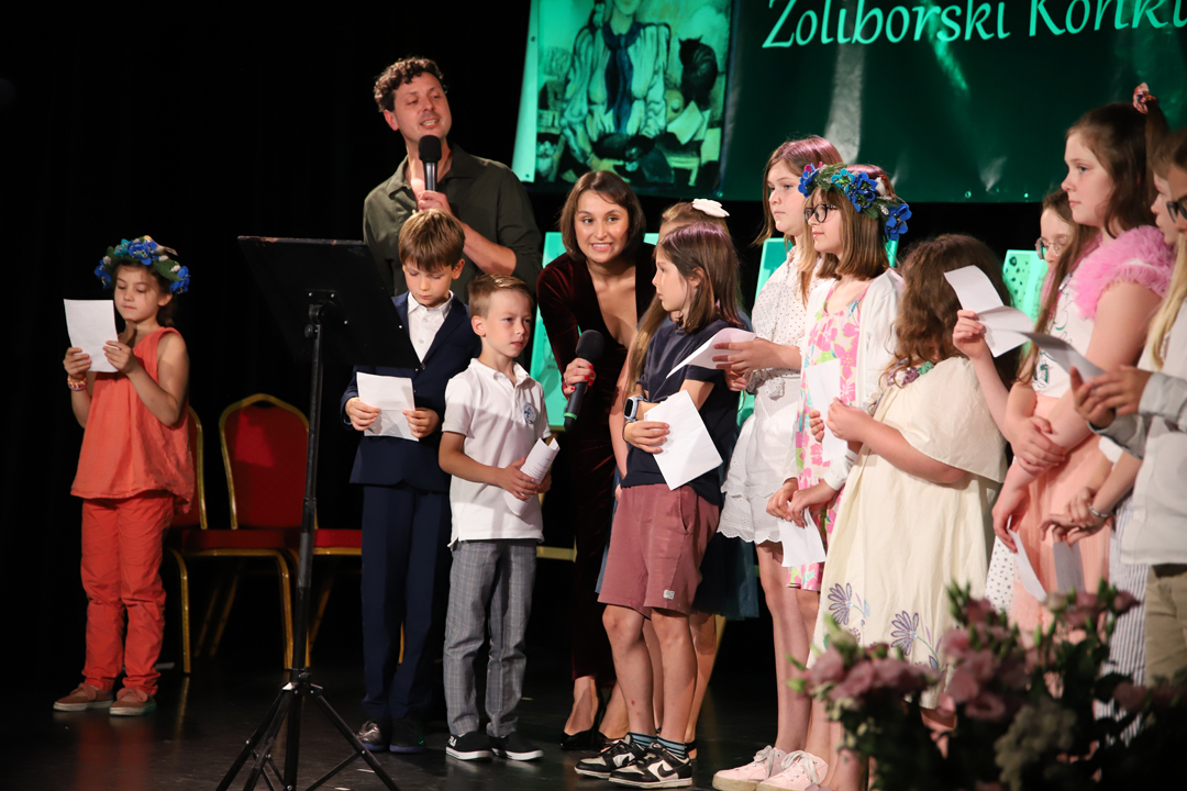 Zdjęcie z II Żoliborskiego Konkursu Recytatorskiego im. Joanny Kulmowej zorganizowanego przez Bibliotekę Publiczną w Dzielnicy Żoliborz, Społeczny Dom Kultury WSM oraz Fundację Joanny i Jana Kulmów. Artyści występujący na scenie Społecznego Domu Kultury śpiewają z laureatami konkursu przy mikrofonach. W tle baner z napisem Żoliborski Konkurs Recytatorski im. Joanny Kulmowej oraz sztalugi z fragmentami utworów Joanny Kulmowej.