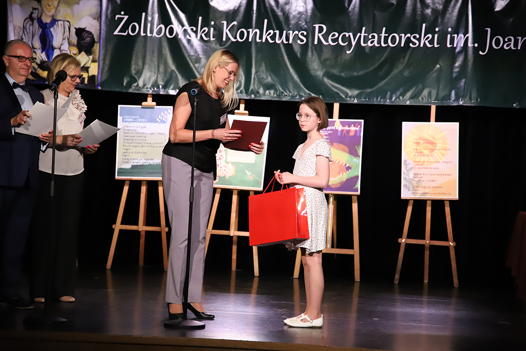 Zdjęcie z II Żoliborskiego Konkursu Recytatorskiego im. Joanny Kulmowej zorganizowanego przez Bibliotekę Publiczną w Dzielnicy Żoliborz, Społeczny Dom Kultury WSM oraz Fundację Joanny i Jana Kulmów. Członek Jury wręcza nagrody i dyplomy na scenie Społecznego Domu Kultury. W tle baner z napisem Żoliborski Konkurs Recytatorski im. Joanny Kulmowej oraz sztalugi z fragmentami utworów Joanny Kulmowej.