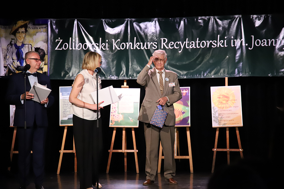 Zdjęcie z II Żoliborskiego Konkursu Recytatorskiego im. Joanny Kulmowej zorganizowanego przez Bibliotekę Publiczną w Dzielnicy Żoliborz, Społeczny Dom Kultury WSM oraz Fundację Joanny i Jana Kulmów. Członek Jury wręcza nagrody i dyplomy na scenie Społecznego Domu Kultury. W tle baner z napisem Żoliborski Konkurs Recytatorski im. Joanny Kulmowej oraz sztalugi z fragmentami utworów Joanny Kulmowej.
