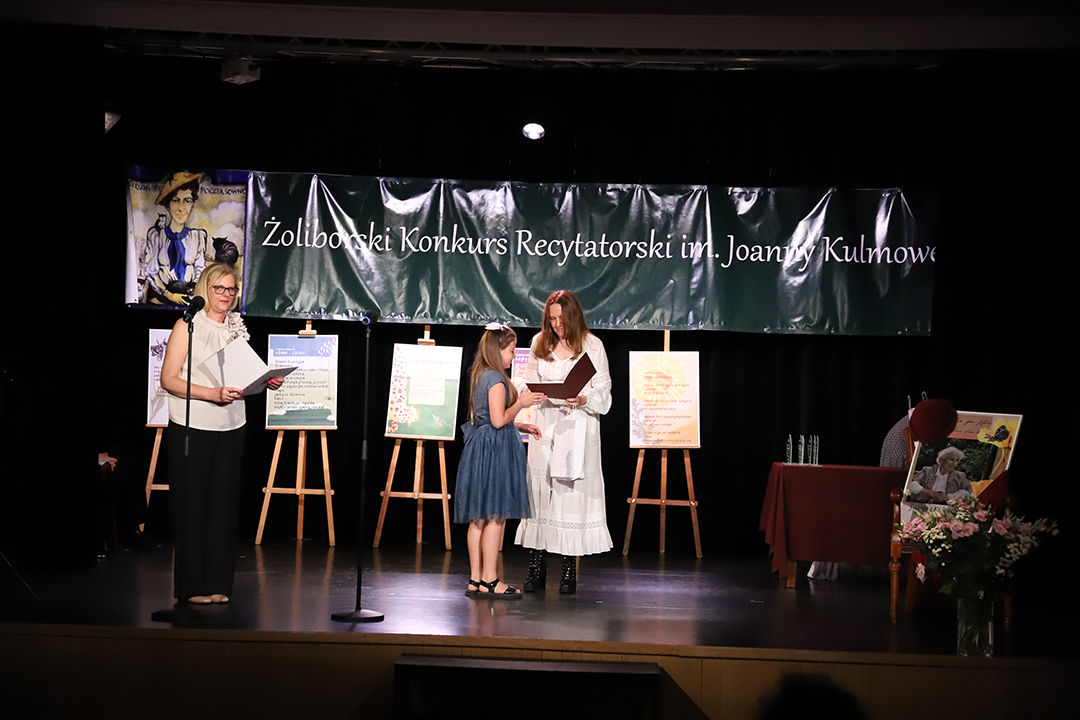 Zdjęcie z II Żoliborskiego Konkursu Recytatorskiego im. Joanny Kulmowej zorganizowanego przez Bibliotekę Publiczną w Dzielnicy Żoliborz, Społeczny Dom Kultury WSM oraz Fundację Joanny i Jana Kulmów. Członek Jury wręcza nagrody i dyplomy na scenie Społecznego Domu Kultury. W tle baner z napisem Żoliborski Konkurs Recytatorski im. Joanny Kulmowej oraz sztalugi z fragmentami utworów Joanny Kulmowej.