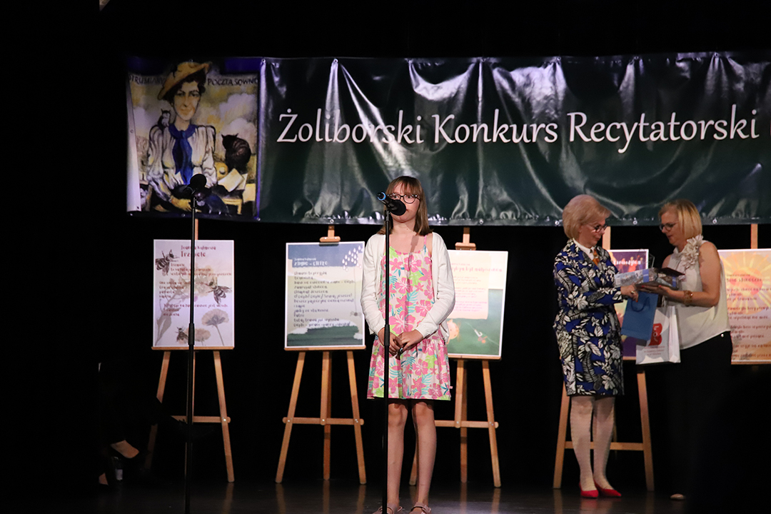 Zdjęcie z II Żoliborskiego Konkursu Recytatorskiego im. Joanny Kulmowej zorganizowanego przez Bibliotekę Publiczną w Dzielnicy Żoliborz, Społeczny Dom Kultury WSM oraz Fundację Joanny i Jana Kulmów. Członek Jury wręcza nagrody i dyplomy na scenie Społecznego Domu Kultury. W tle baner z napisem Żoliborski Konkurs Recytatorski im. Joanny Kulmowej oraz sztalugi z fragmentami utworów Joanny Kulmowej.