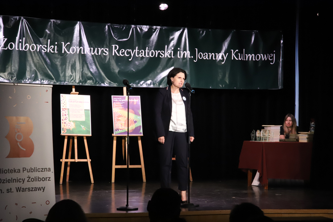Zdjęcie z II Żoliborskiego Konkursu Recytatorskiego im. Joanny Kulmowej zorganizowanego przez Bibliotekę Publiczną w Dzielnicy Żoliborz, Społeczny Dom Kultury WSM oraz Fundację Joanny i Jana Kulmów. Burmistrzyni Żoliborza przemawia na scenie do mikrofonu. W tle baner z napisem Żoliborski Konkurs Recytatorski im. Joanny Kulmowej oraz sztalugi z fragmentami utworów Joanny Kulmowej oraz rollupem Biblioteki Żoliborskiej z napisem Biblioteka Publiczna w Dzielnicy Żoliborz.