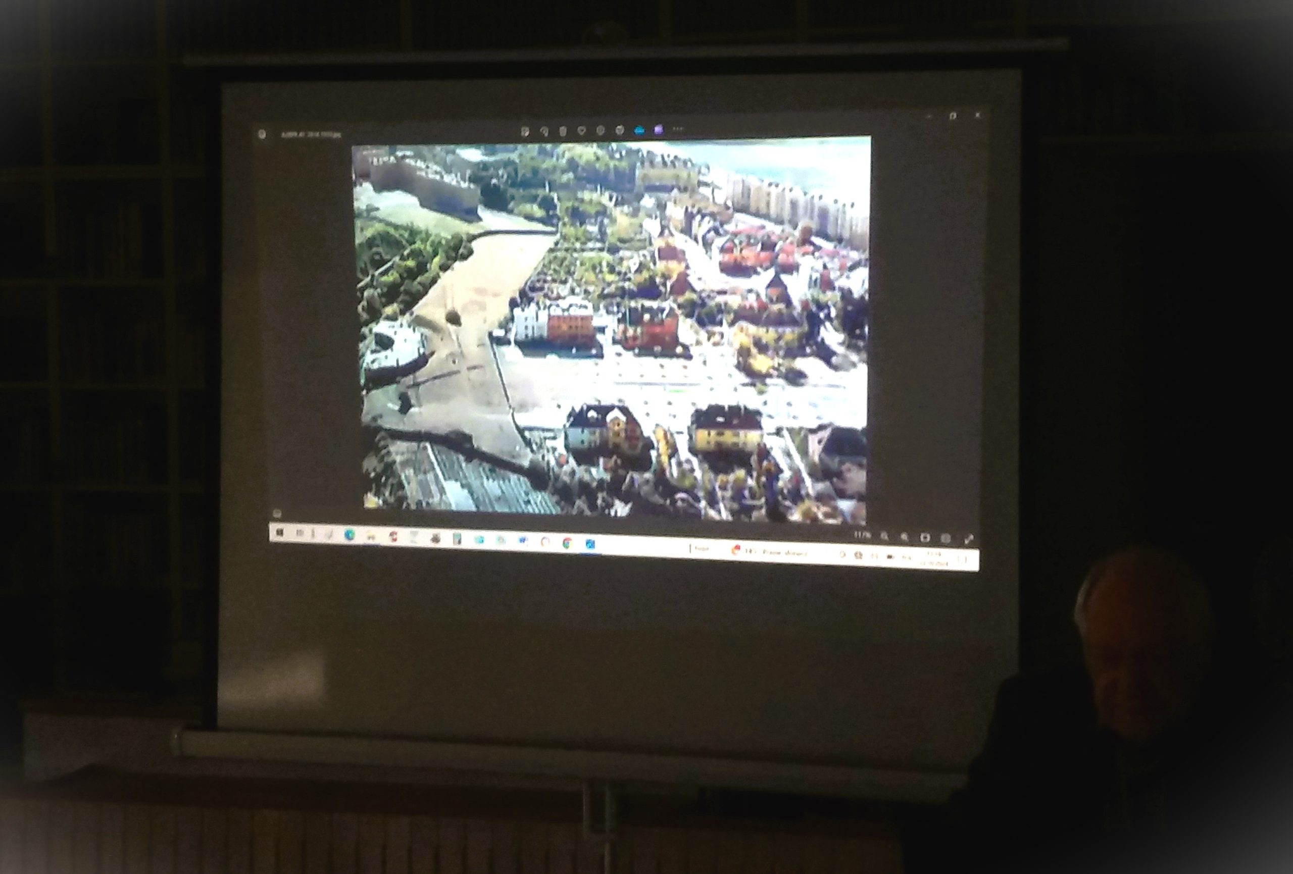 Zdjęcie przedstawia wnętrze biblioteki. Na tablicy stojącej w centrum widać stare zdjęcie. Obok tablicy siedzi mężczyzna. Na pierwszym planie widać ludzi.