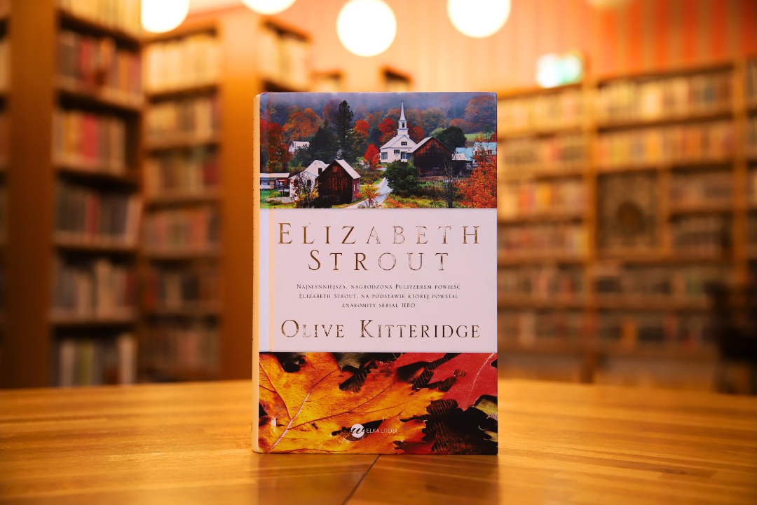 zdjecie przedstawia okładkę książki olive kitteridge elizabeth strout na tle wnętrza wypożyczalni nr 16, widoczne napisy: elizabeth strout najsłynniejsza nagrodzona pulitzerem powieść elizabeth strout, na podstawie której powstał znakomity serila hbo, olive kitteridge