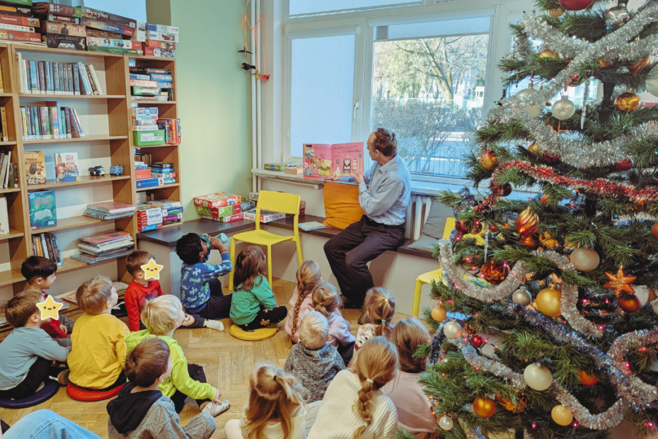 zdjęcie mężczyzny siedzącego i czytającego książkę siedzącym na podłodze dzieciom we wnętrzu biblioteki dla dzieci nr xv. widać choinkę ubraną w bombki, łańcuchy i światełka oraz regały biblioteczne.