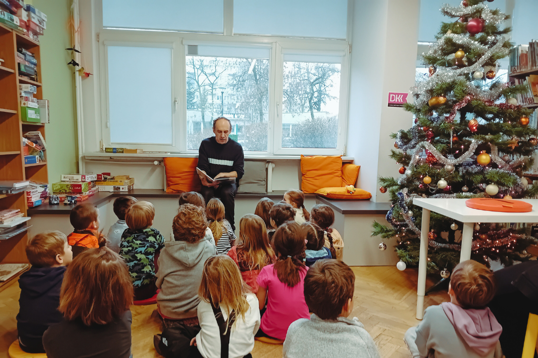 zdjęcie mężczyzny siedzącego i czytającego książkę siedzącym na podłodze dzieciom we wnętrzu biblioteki dla dzieci nr xv. widać choinkę ubraną w bombki, łańcuchy i światełka oraz regały biblioteczne.