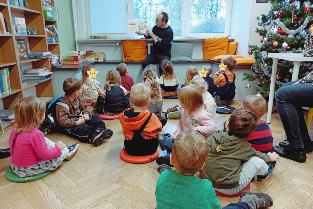 zdjęcie mężczyzny siedzącego i czytającego książkę siedzącym na podłodze dzieciom we wnętrzu biblioteki dla dzieci nr xv. widać choinkę ubraną w bombki, łańcuchy i światełka oraz regały biblioteczne.