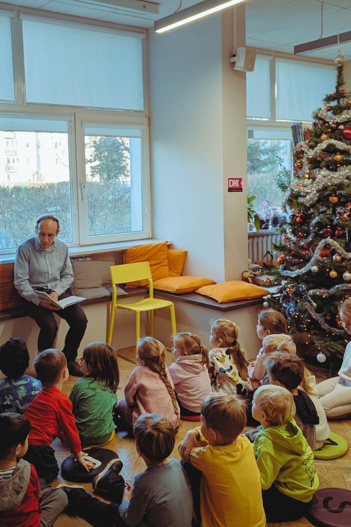 zdjęcie mężczyzny siedzącego i czytającego książkę siedzącym na podłodze dzieciom we wnętrzu biblioteki dla dzieci nr xv. widać choinkę ubraną w bombki, łańcuchy i światełka oraz regały biblioteczne.