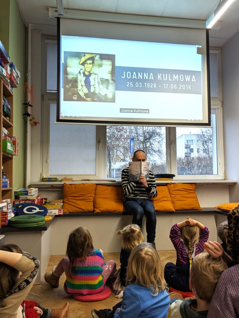 mężczyzna siedzący pod rzutnikiem z planszą z ilustracją Joanny Kulmowej oraz napisem Joanna Kulmowa 25.03.1928-17.06.2018 zakrywa twarz książką echa śpiące przed siedzącymi przed nim dziećmi we wnętrzu biblioteki dla dzieci nr xv