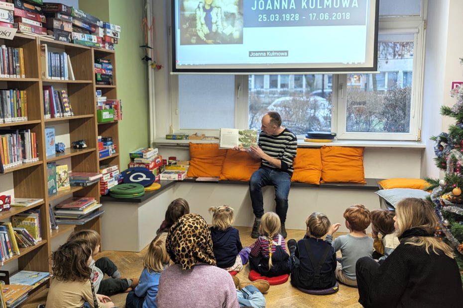 mężczyzna siedzący pod rzutnikiem z planszą z ilustracją Joanny Kulmowej oraz napisem Joanna Kulmowa 25.03.1928-17.06.2018 czyta książkę Kolory Joanny Kulmowej do siedzących przed nim dziećmi we wnętrzu biblioteki dla dzieci nr xv