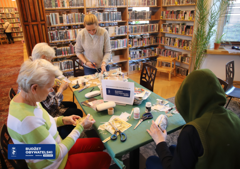 Zdjęcie z warsztatów "Rodzinne Warsztaty Artystyczne - Ozdabiamy etui na okulary oraz świeczniki metodą decoupage" zorganizowanego przez Bibliotekę Publiczną w Dzielnicy Żoliborz 23 kwietnia 2024 roku. Uczestniczki warsztatów dekorujące etui na okulary oraz świeczniki metodą decoupage. Tabliczka ze znaczkiem budżetu obywatelskiego z napisami: Budżet Obywatelski Zgłaszam Głosuję Korzystam na stole pośród przyrządów do dekorowania metodą decoupage. W lewym dolnym rogu znaczek budżetu obywatelskiego z napisem: Budżet Obywatelski Zgłaszam Głosuję Korzystam