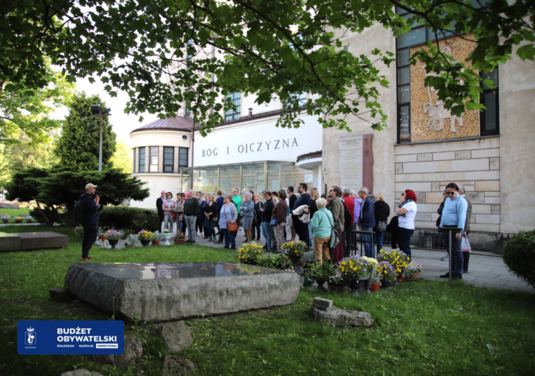 Zdjęcie ze spaceru Żoliborz Urzędniczy zorganizowanego przez Bibliotekę Publiczną w Dzielnicy Żoliborz 8 maja 2024 roku. Przewodnik i grupa słuchaczy stojących przy grobie ks. Jerzego Popiełuszki przy Kościele św. Stanisława Kostki W lewym dolnym rogu znaczek budżetu obywatelskiego z napisem: Budżet Obywatelski Zgłaszam Głosuję Korzystam