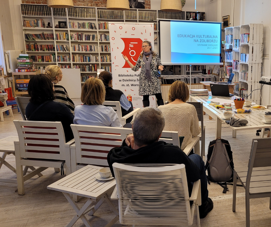 Zdjęcie przedstawia spotkanie dotyczące edukacji kulturalnej. Na pierwszym planie ustawione białe stoliki i krzesła, na których siedzi odwrócone plecami 6 osób (5 kobiet i jeden mężczyzna). Trochę dalej stoi odwrócona twarzą kobieta za nią wyświetlona prezentacja z tekstem: Edukacja kulturalna na Żoliborzu. Spotkanie sieciujące. Obok baner z tekstem Biblioteka Publiczna w dzielnicy Żoliborz m.st. Warszawy.