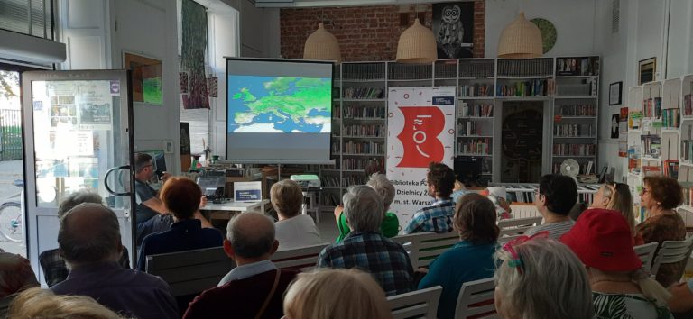 grupa ludzi siedzi i ogląda wyświetlaną na ekranie prezentację, w tle logo biblioteki publicznej oraz logo budżetu obywatelskiego