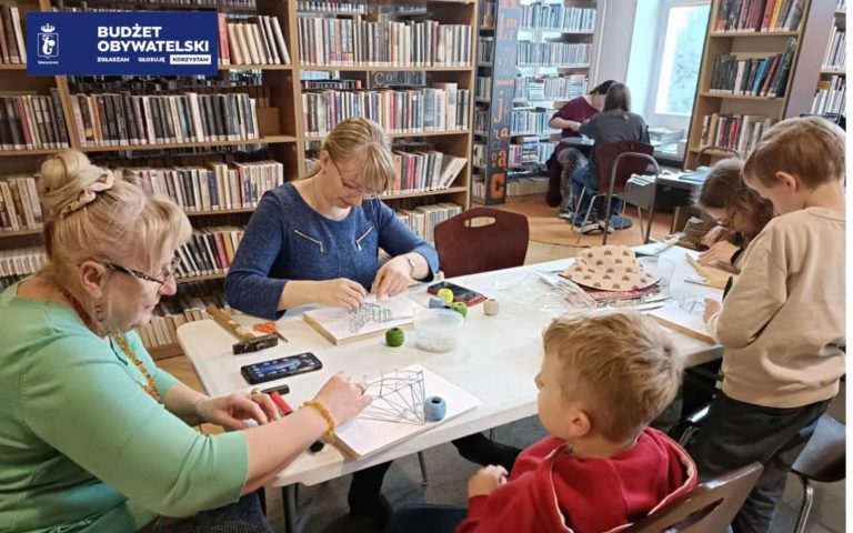 Uczestnicy warsztatów String Art. Na pierwszym planie - Siedzący przy stole warsztatowym trzy kobiety w różnym wieku oraz dwójka dzieci. W tle mniejszy stolik warsztatowy z 2 uczestników zajęć. Uczestnicy zajęć przy pomocy młotków wbijają w drewnianą deseczkę gwoździe, na których rozciągają kolorowe nitki, tworząc w taki sposób przestrzenne obrazy.