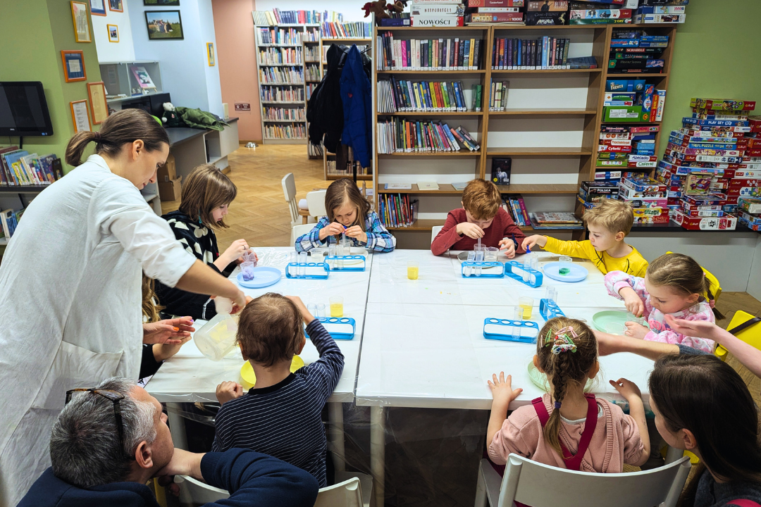 zdjecie z warsztatów "to nie magia, to nauka - "eksperymenty zimowe" we wnętrzu wypożyczalni nr 138 i biblioteki dla dzieci nr XV przy ul. Broniewskiego 9a ósemka dzieci oraz dwoje dorosłych wokół stołu z eksperymentami z wykorzystaniem różnych substancji, pojemników, próbówek i pipetek