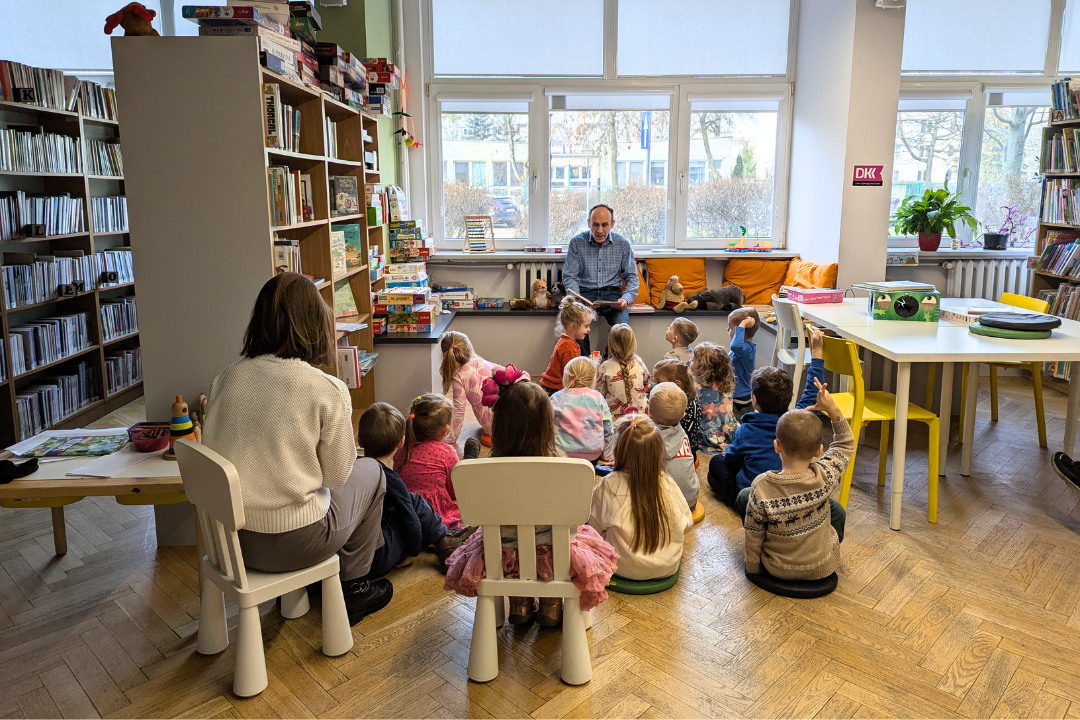 zdjęcie mężczyzny siedzącego i czytającego książkę siedzącym na podłodze i na krzesełkach dzieciom we wnętrzu biblioteki dla dzieci nr xv. w tle regały biblioteczne.