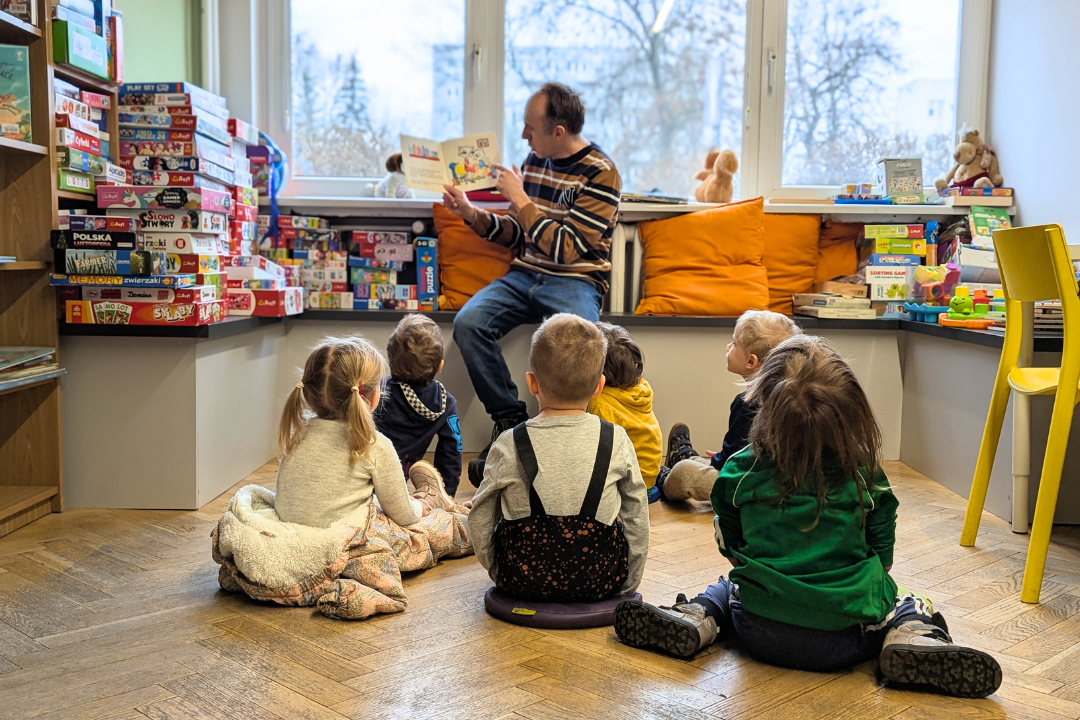 zdjęcie mężczyzny siedzącego i czytającego książkę siedzącym na podłodze dzieciom we wnętrzu biblioteki dla dzieci nr xv. w tle regały biblioteczne.