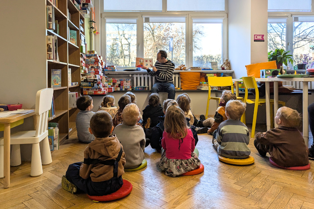 zdjęcie mężczyzny siedzącego i czytającego książkę siedzącym na podłodze dzieciom we wnętrzu biblioteki dla dzieci nr xv. w tle regały biblioteczne.