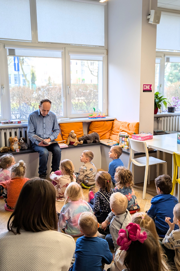 zdjęcie mężczyzny siedzącego i czytającego książkę siedzącym na podłodze dzieciom we wnętrzu biblioteki dla dzieci nr xv. w tle regały biblioteczne.