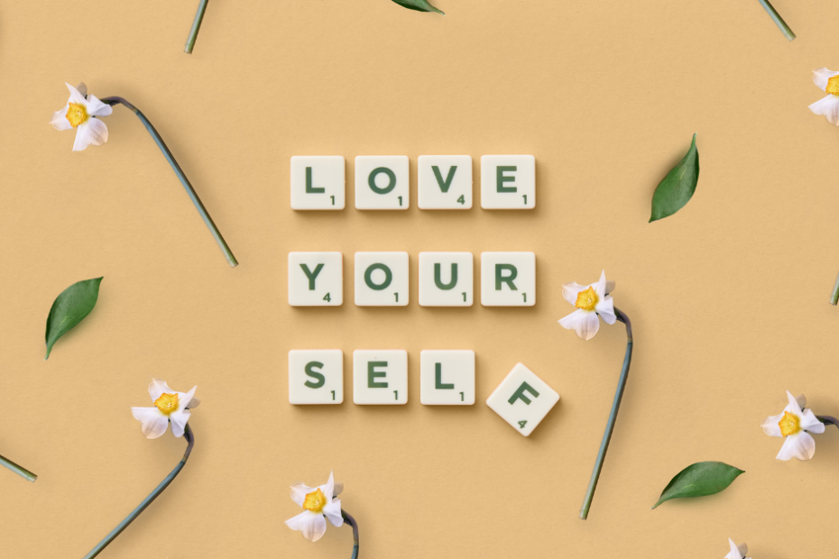 Na żółtym tle rozłożone zielone liście i kwiaty narcyzów oraz napis z klocków do gry scrabble: Love yourself