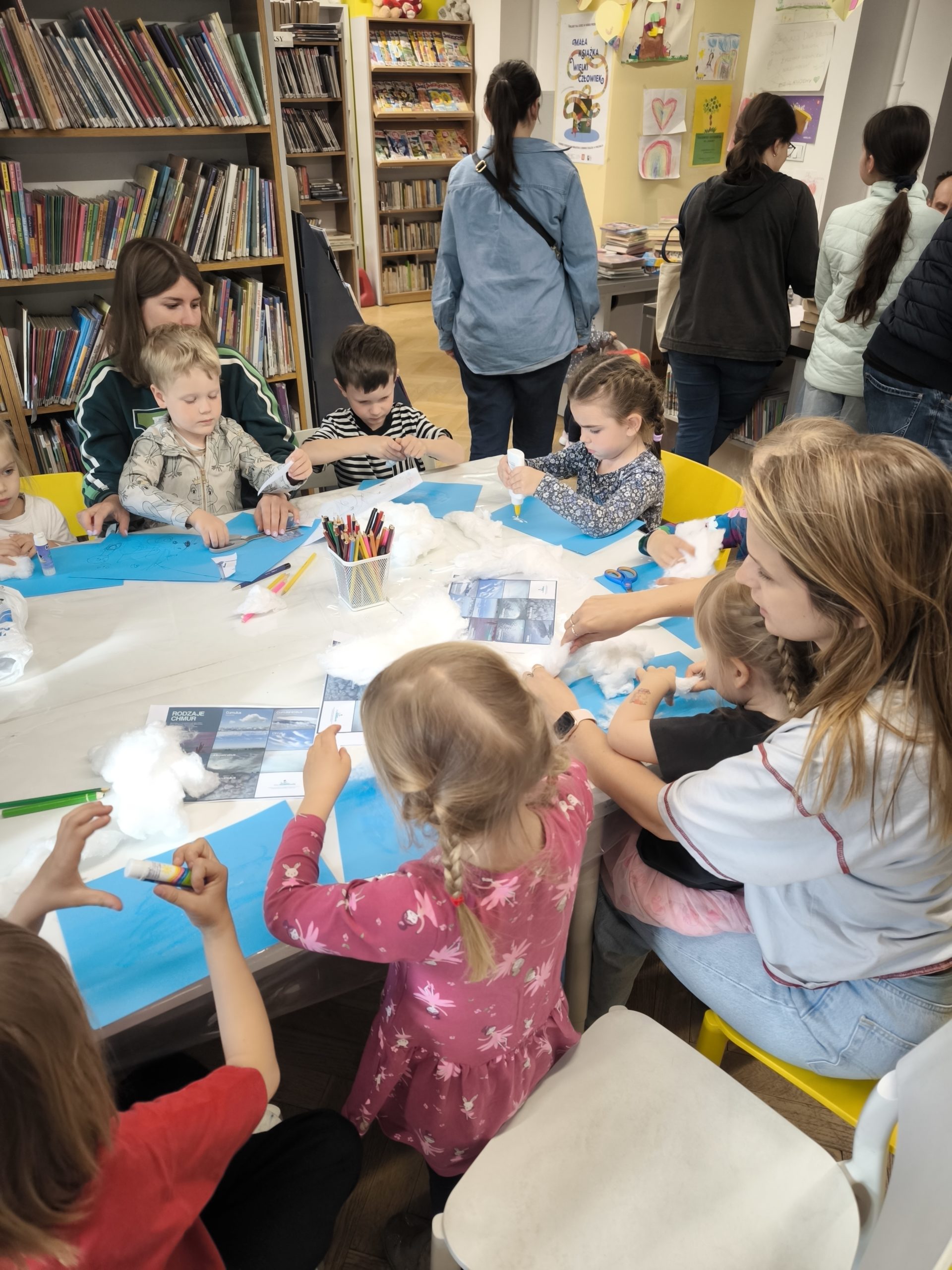zdjecie z warsztatów "to nie magia, to nauka - wiosenne slajmy" we wnętrzu wypożyczalni nr 138 i biblioteki dla dzieci nr XV przy ul. Broniewskiego 9a. grupa dzieci, które wykonują eksperymenty przy stole