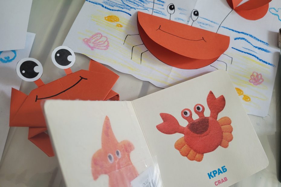 Na białym stole czerwone papierowe kraby, kartki białego papieru z rysunkami oraz mała książeczka dla dzieci z krabem i tekstem krab po angielsku i ukraińsku.