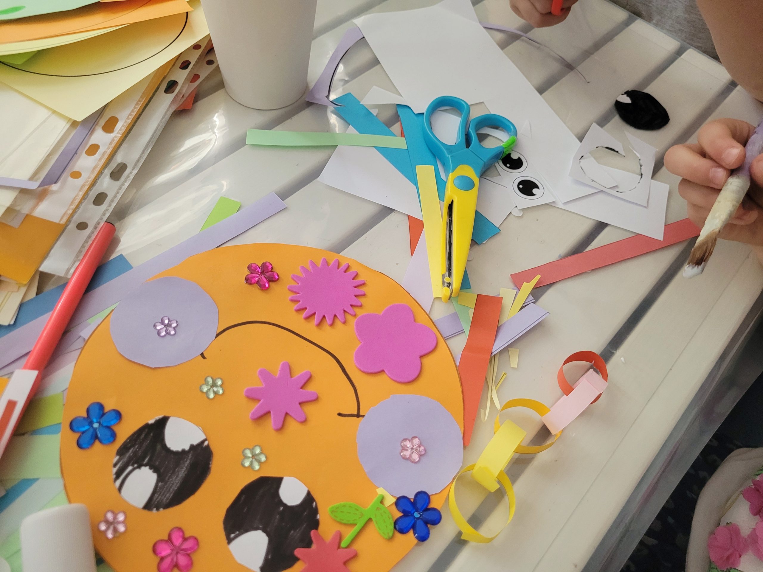 Przy stole kilka osób (widać tylko ręce) wykonują papierowe ośmiornice na stole materiały plastyczne: flamastry, papier, klej, nożyce