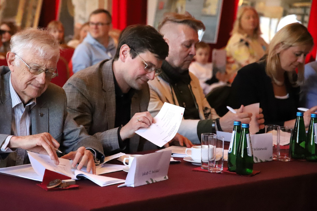 Zdjęcie z III Żoliborskiego Konkursu Recytatorskiego im. Joanny Kulmowej zorganizowanego przez Bibliotekę Publiczną w Dzielnicy Żoliborz, Społeczny Dom Kultury WSM oraz Fundację Joanny i Jana Kulmów. Członkowie Jury podczas przesłuchania. 4 osoby siedzące przy stole i przeglądające kartki.