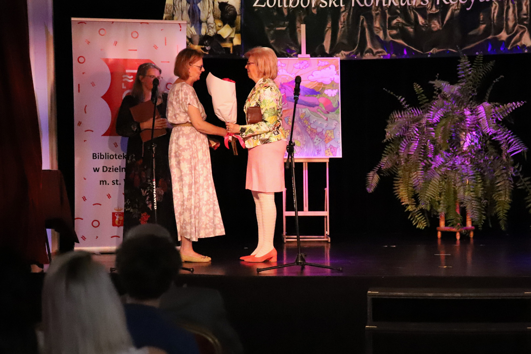 Zdjęcie z III Żoliborskiego Konkursu Recytatorskiego im. Joanny Kulmowej zorganizowanego przez Bibliotekę Publiczną w Dzielnicy Żoliborz, Społeczny Dom Kultury WSM oraz Fundację Joanny i Jana Kulmów. 3 kobiety na scenie, jedna wręczająca drugiej kwiaty.